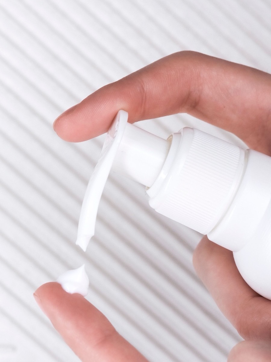 A hand pressing the pump of a plain unlabeled white bottle, dispensing a little lotion onto a fingertip against a white ribbed background