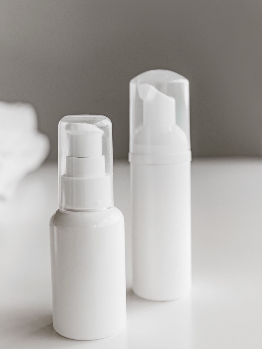 An unlabeled white pump bottle and a white foam-pump bottle on a white table, with a white towel in the background